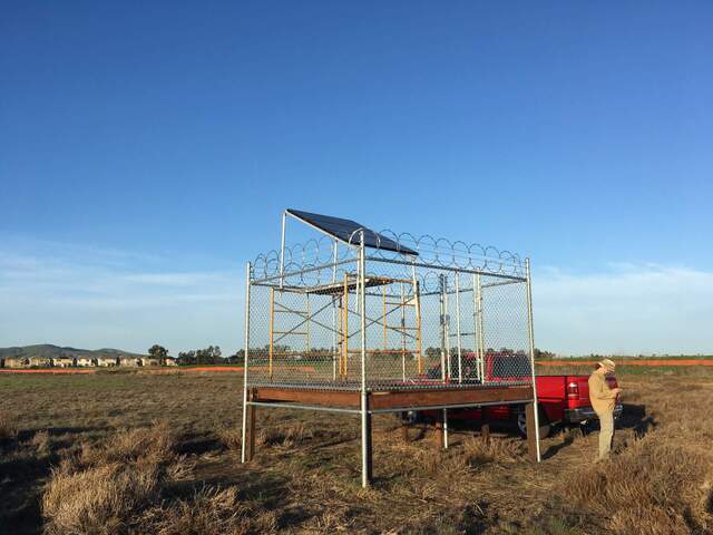 Solar panels set up on scaffolding inside security fence. The bottom panel is damaged and needs to be replaced. Suisin City houses are in the background.